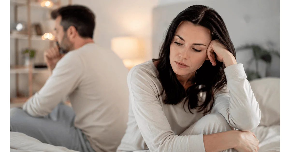 Couple sitting apart in a bedroom, reflecting the emotional distance and personal distress associated with a persistent lack of sexual interest.